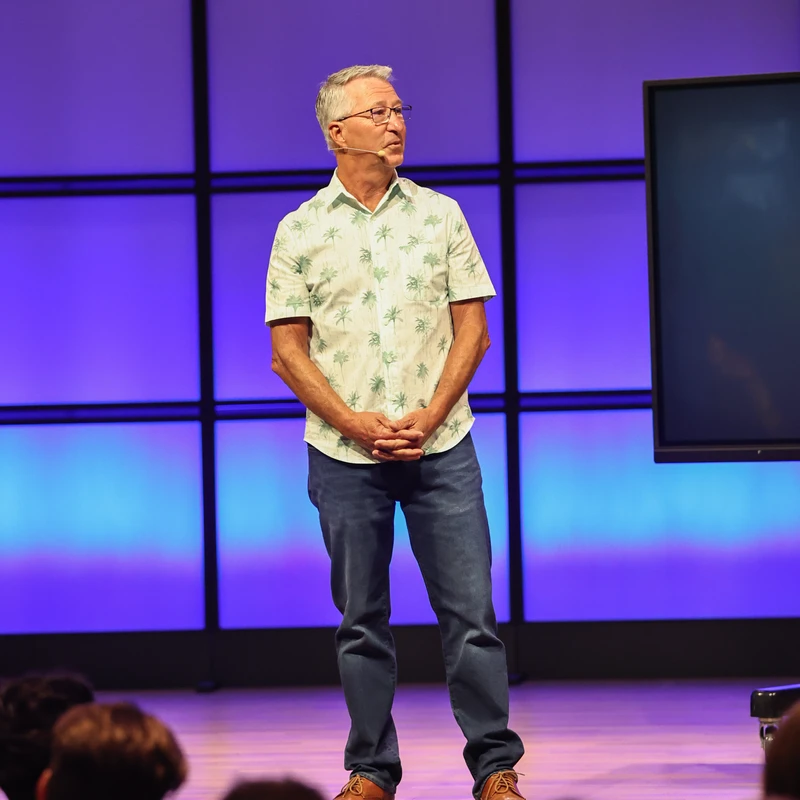 An older man with glasses stands on a stage in front of a blue and purple backdrop, wearing a palm tree shirt and jeans while addressing an audience.