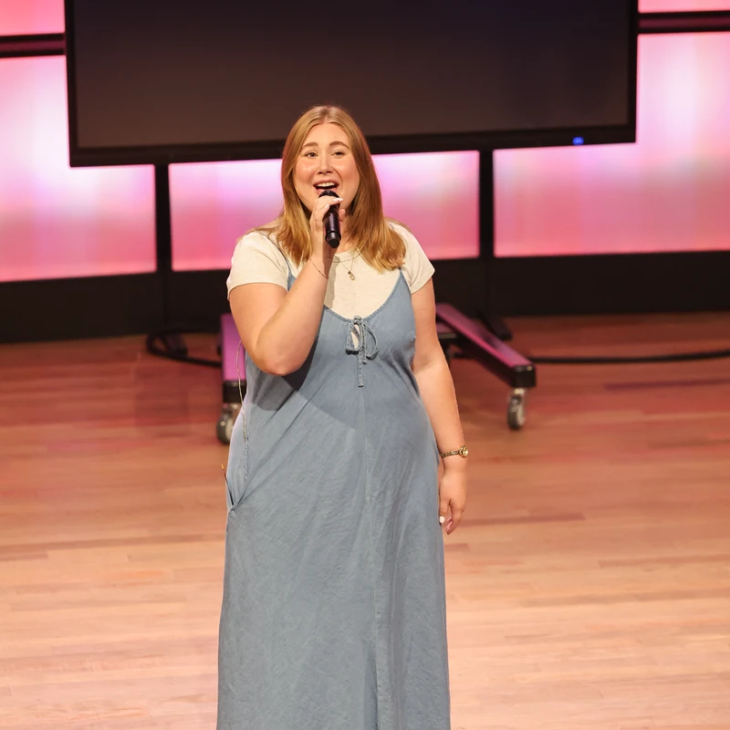A young woman with long blonde hair sings into a microphone while standing on a wooden stage. She wears a blue denim jumper over a white t-shirt.