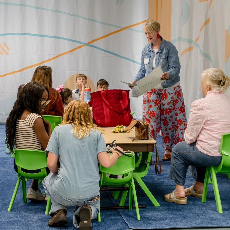 A group of adults and children sit at a table while a woman in a denim jacket stands and speaks to them in a brightly decorated classroom setting.