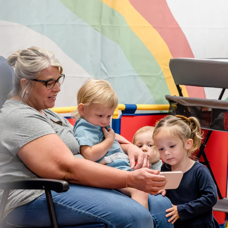 A woman wearing glasses sits in a chair with a toddler on her lap, showing them and two other young children something on a smartphone.