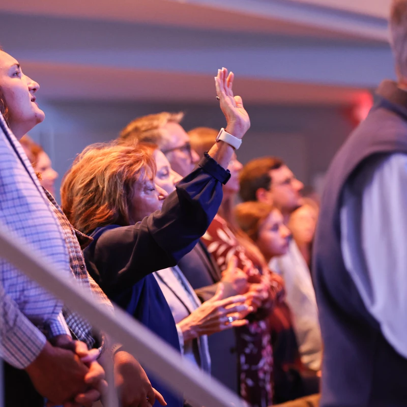 A diverse group of people stand together in a dimly lit auditorium. A woman in the center has her hand raised in a gesture of praise or worship.