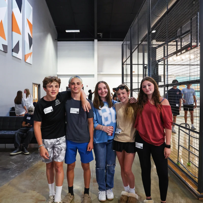 Five teenagers, two boys and three girls, stand smiling for a photo in a long hallway, with their arms around each other next to a caged sports court.