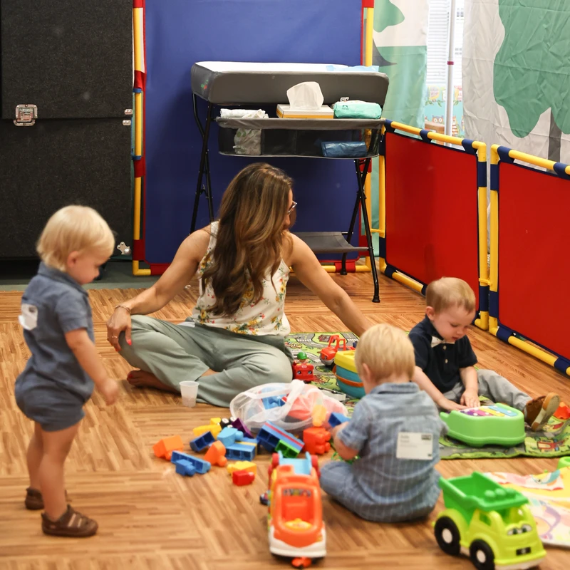 A woman sits on a wood-patterned floor with three toddlers in a playroom. They are surrounded by colorful toys like blocks and trucks.