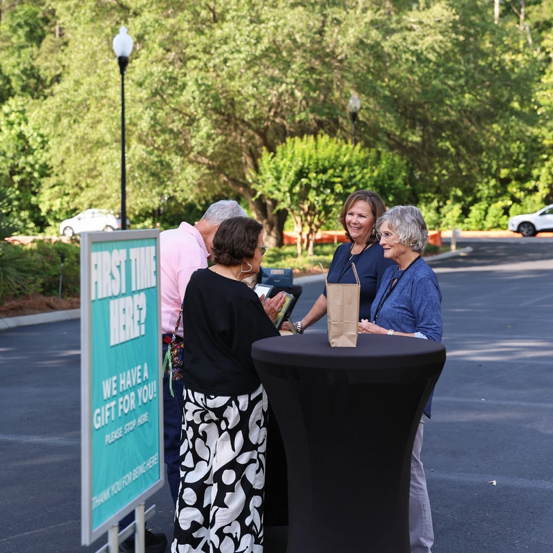 Four people gather around a tall black table at a welcome station. A sign reads, 'First Time Here? We have a gift for you!' in an outdoor setting.