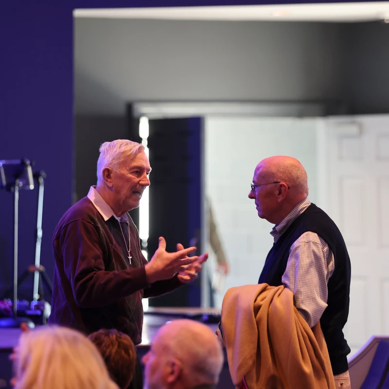 Two older men stand talking in a dimly lit auditorium. The man on the left, wearing a cross necklace, gestures with his hands as he speaks to the other man.