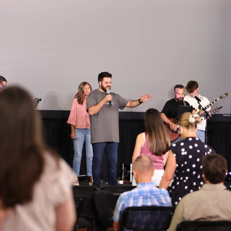 A bearded man in a grey t-shirt stands on a stage, speaking into a microphone. A band is behind him and an audience is seated in front of him.