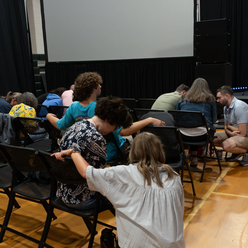 A group of people sit on folding chairs in a gym facing a large screen. Some are kneeling or bowing their heads in prayer, with hands on each other's shoulders.