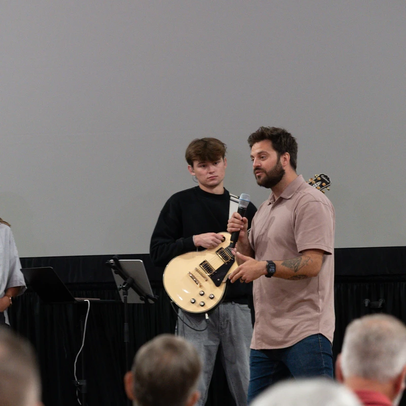 A man with a beard speaks into a microphone on stage. To his left, a young man holds an electric guitar and a woman watches, with an audience in front.