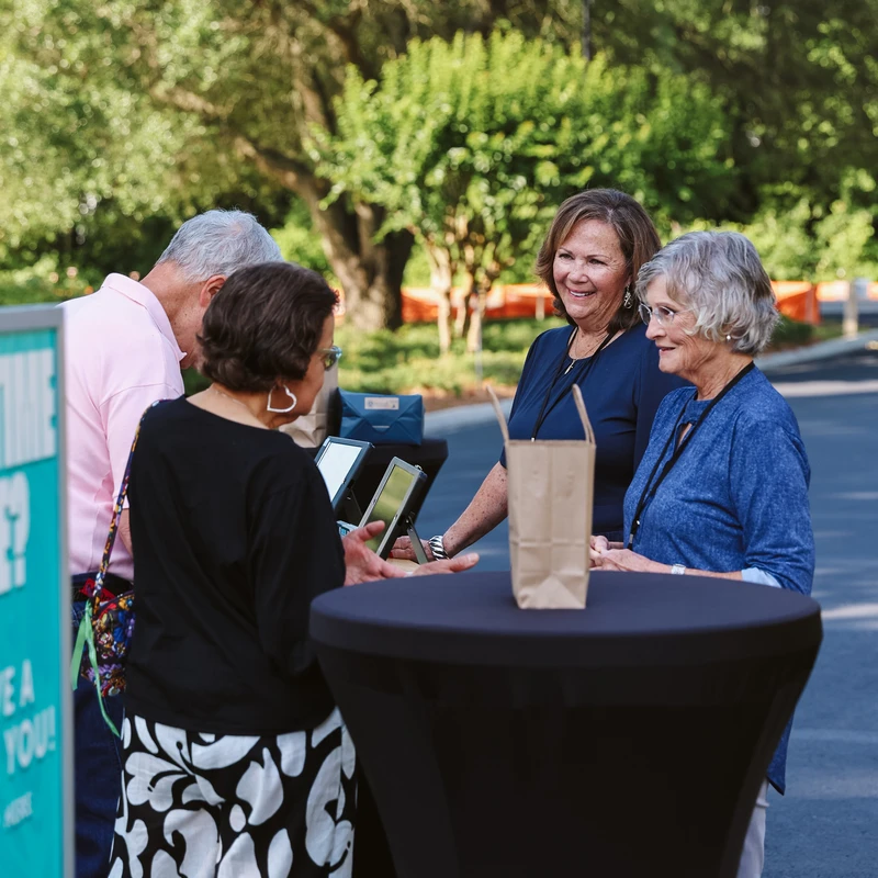 Four older adults stand around a tall black table at an outdoor event, smiling and talking near a check-in station with tablets.