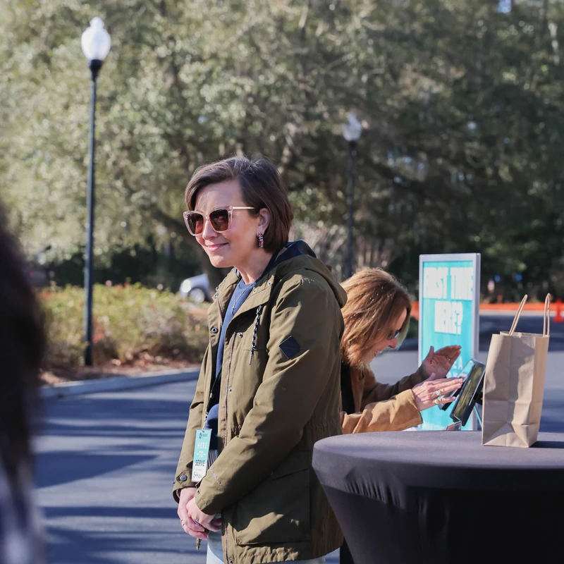 A smiling woman with short brown hair and sunglasses, wearing a green jacket and a 'Here to Serve' lanyard, stands outdoors at a welcome event.