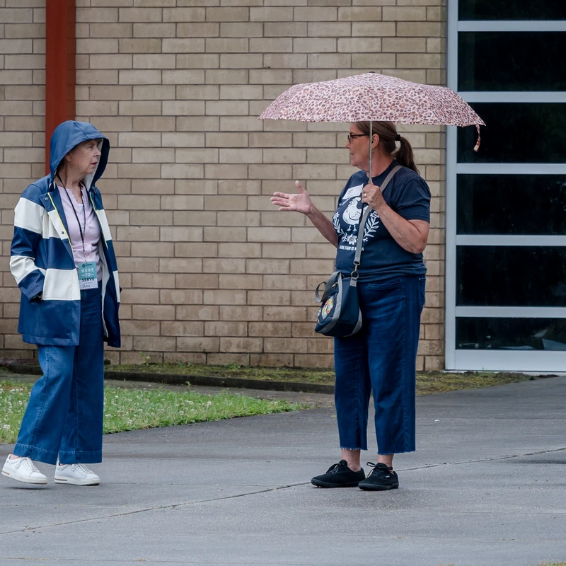 Two women stand on a sidewalk talking. One wears a blue and white hooded raincoat, while the other holds a leopard-print umbrella and gestures as she speaks.