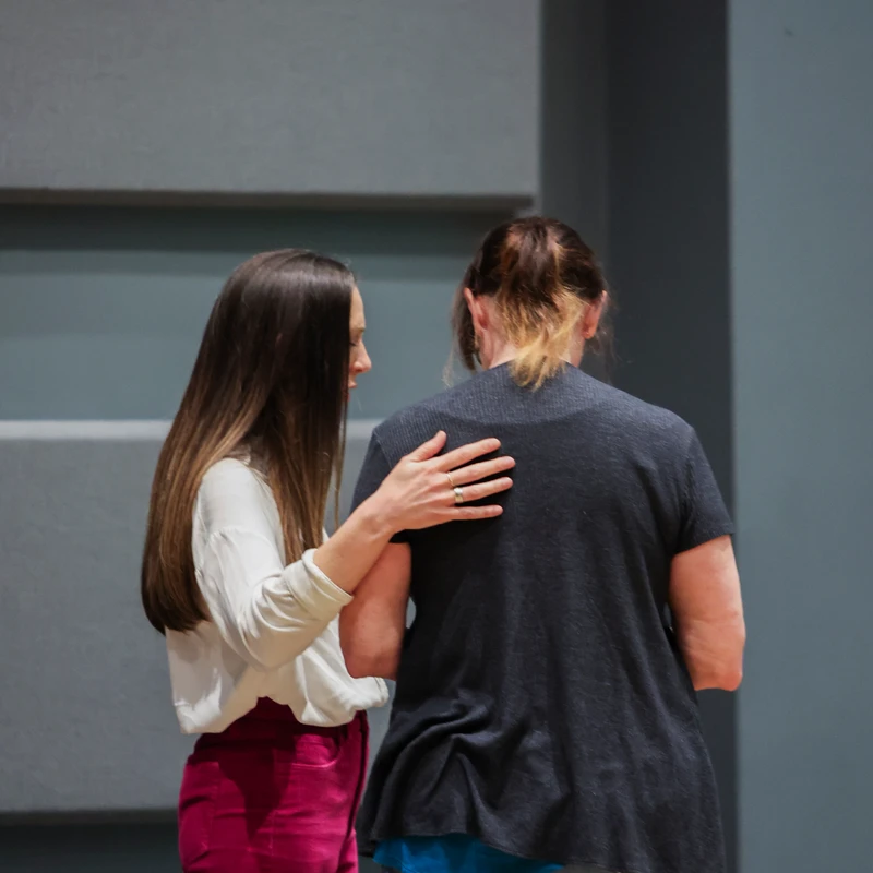 A woman with long dark hair places a supportive hand on the back of another woman who is facing away from the camera, against a grey paneled wall.