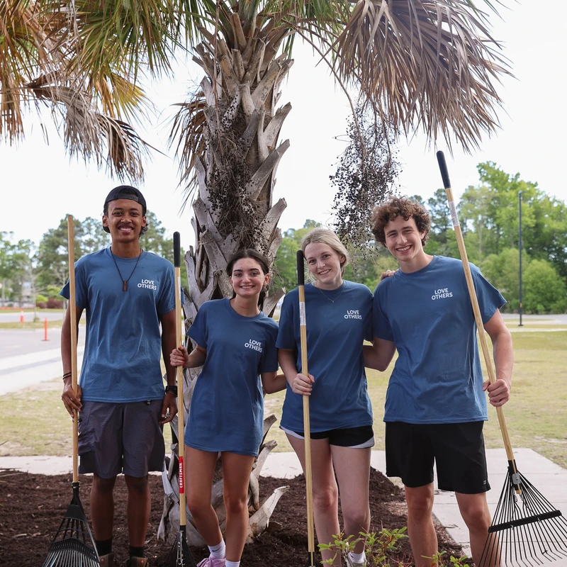 Four young volunteers in matching blue 'Love Others' shirts smile for a photo while holding rakes. They are standing on fresh mulch around a palm tree.