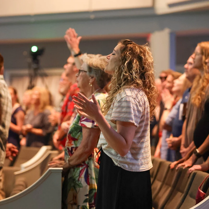 A young woman with curly hair stands with eyes closed and hands raised in worship, surrounded by a congregation in a church sanctuary.