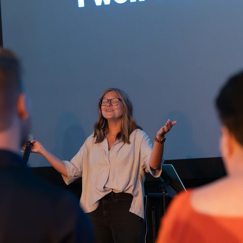 A woman with glasses sings on stage, holding a microphone with arms open. Behind her, a large screen displays song lyrics.