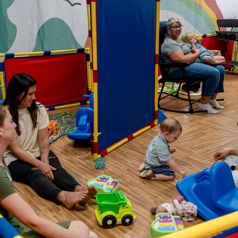 A group of women and toddlers interact in a colorful playroom. Some women sit on the floor chatting, while a toddler plays with toys nearby.