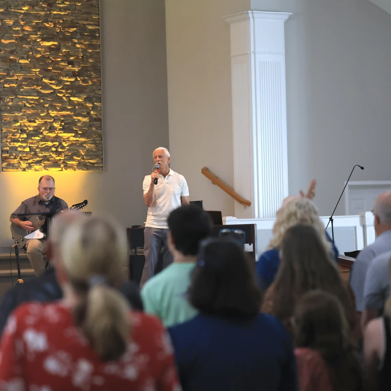 A man in a white polo shirt sings into a microphone, leading a church service, with a guitarist beside him and the congregation facing them.