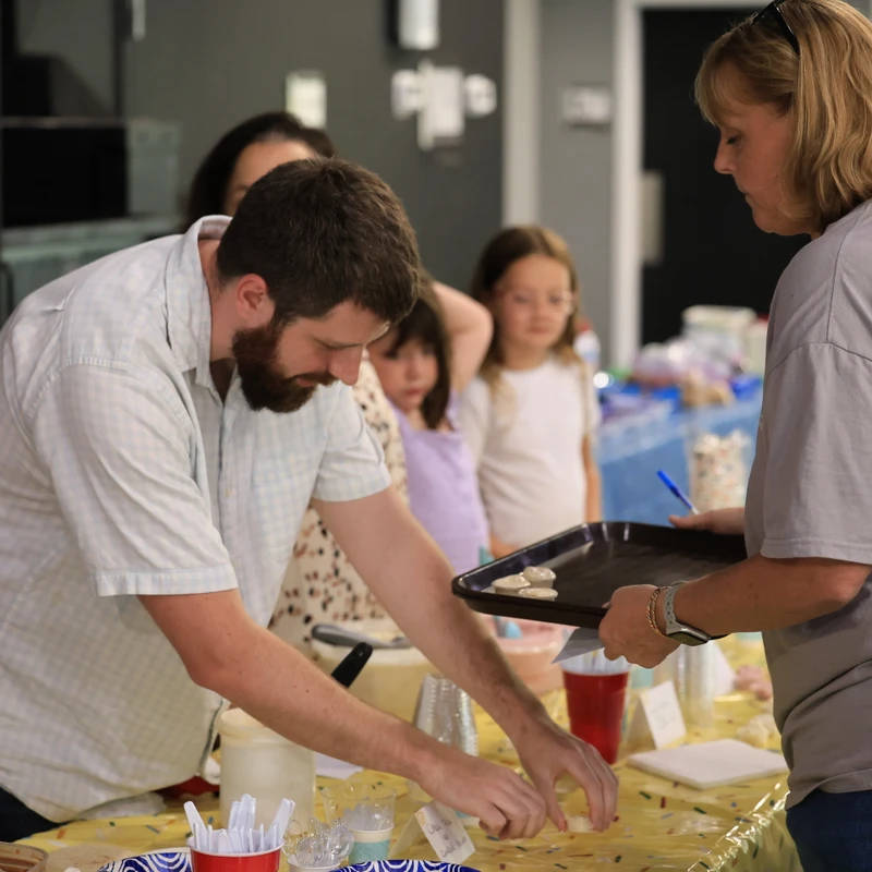 A man with a beard prepares food on a table while a woman holds a tray. Two young girls watch them from the background at an indoor gathering.