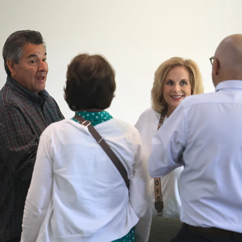 Four adults stand together against a plain white wall, talking. A man in a plaid shirt speaks, and a blonde woman in the middle smiles at the group.