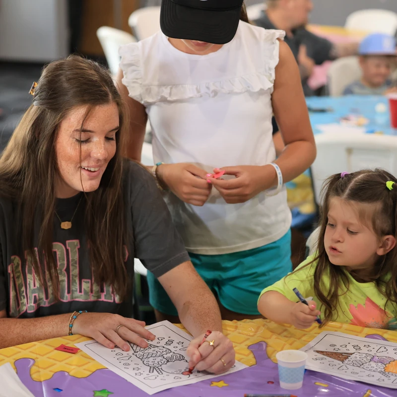 A smiling young woman and two young girls sit at a colorful table, focused on coloring pictures of ice cream cones together during an event.