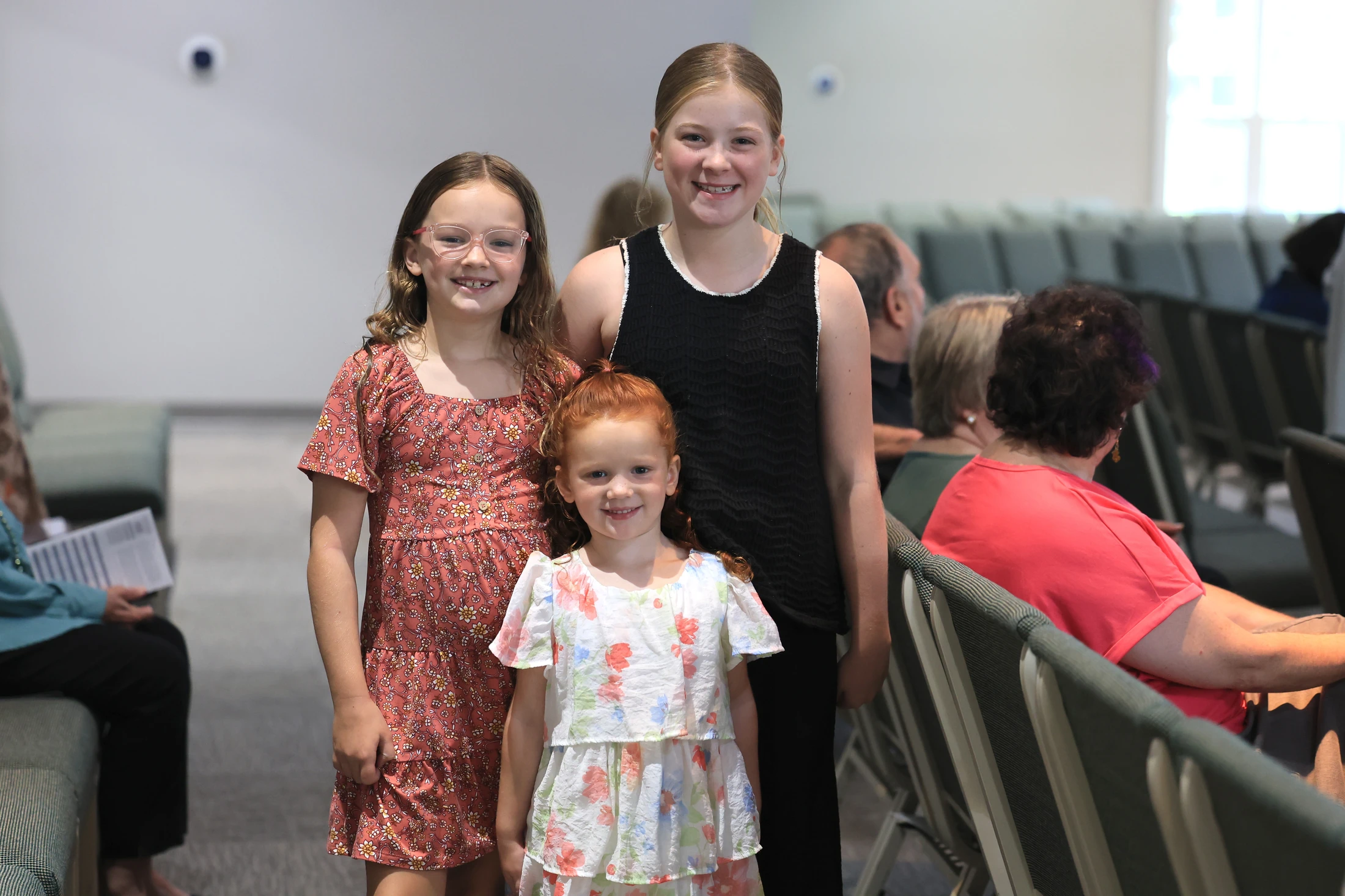 Three young girls smile for the camera while standing in an aisle of a large room with rows of seating. A redhead stands in front of two older girls.