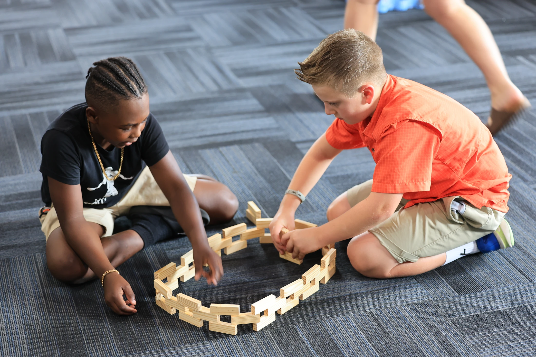 Two young boys, one Black and one white, sit on a grey carpet working together to build a circular structure with rectangular wooden blocks.