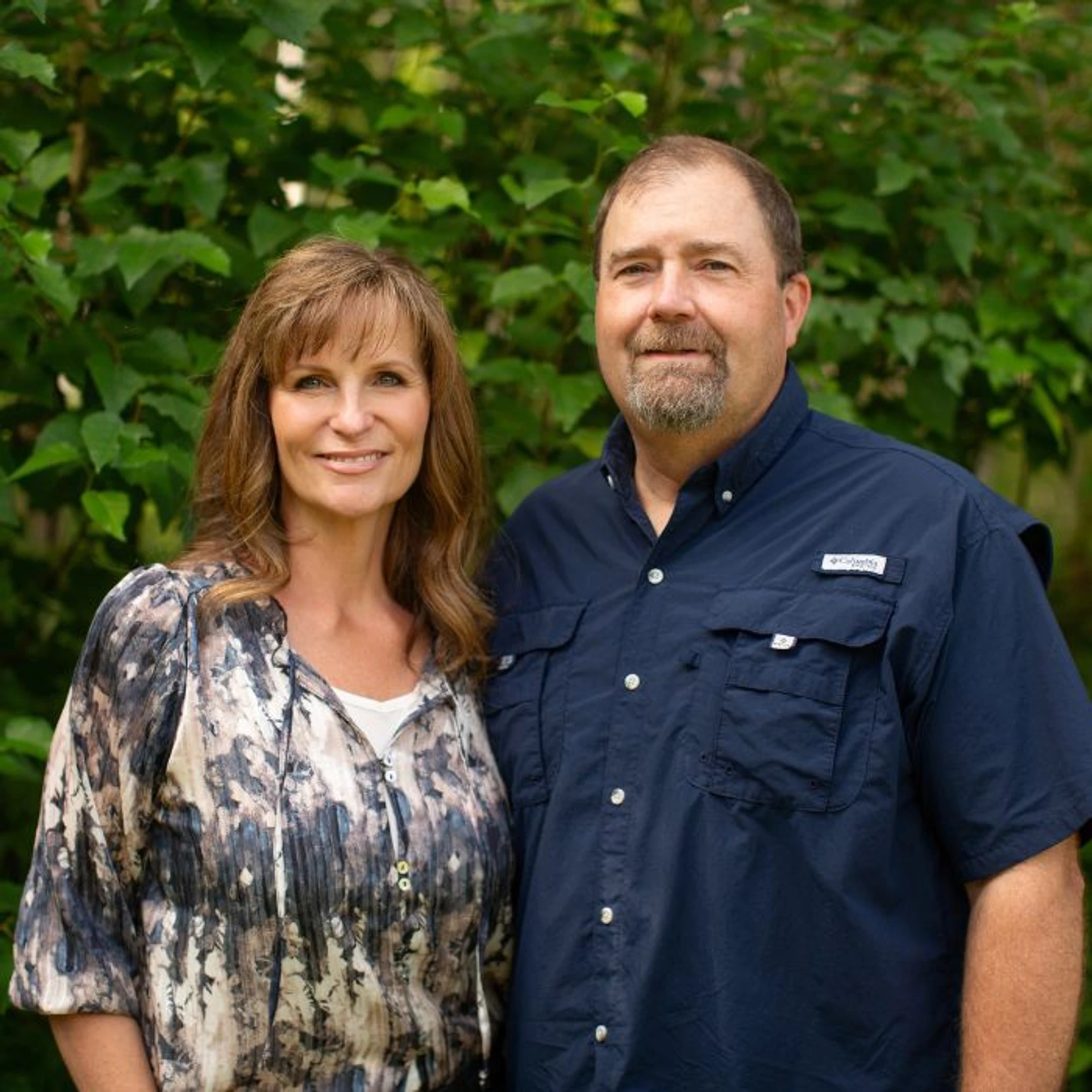A man and a woman stand side-by-side outdoors in front of green leaves. The woman has brown hair and wears a patterned blouse. The man wears a blue shirt.
