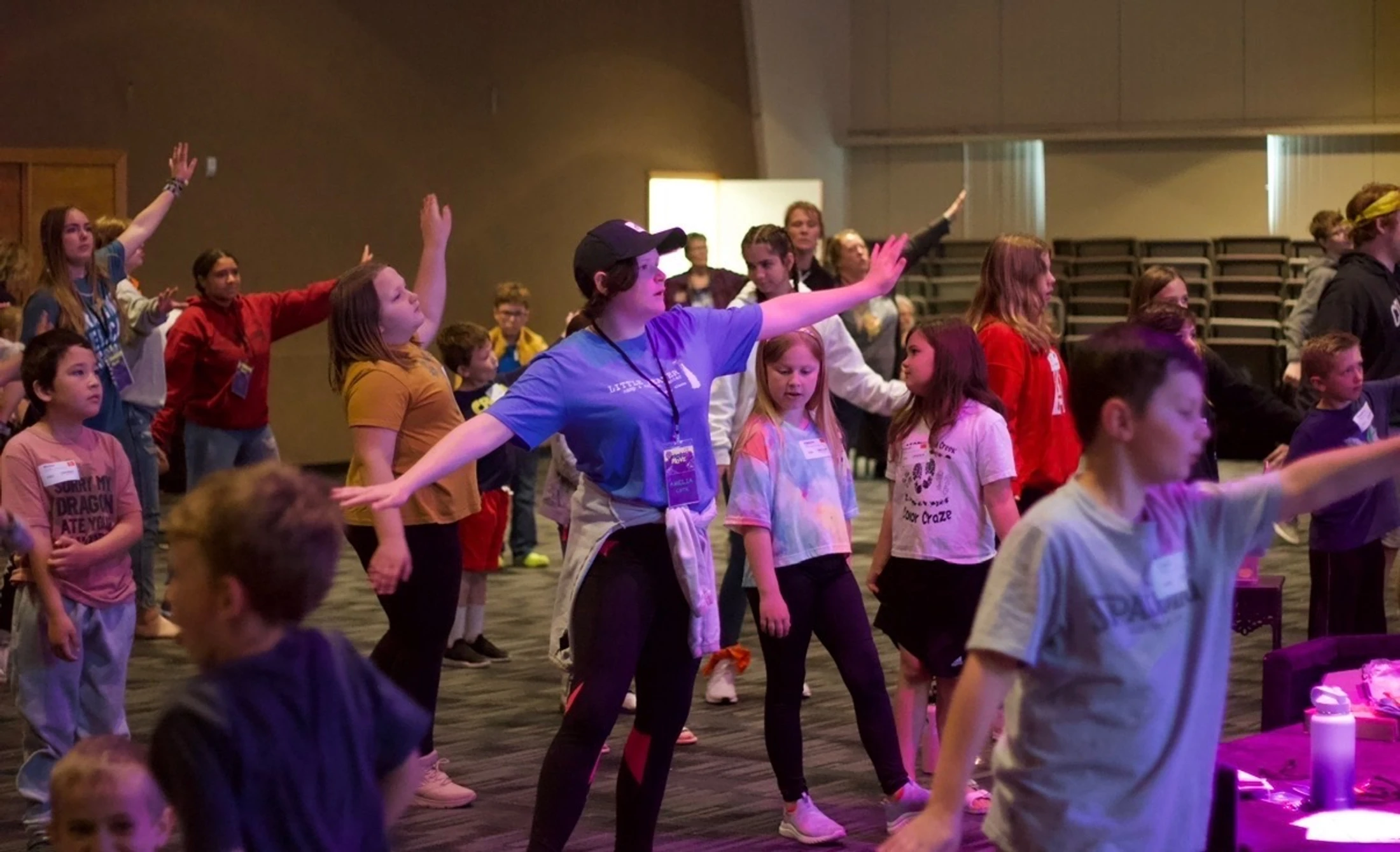A group of children and adults follow a leader in a dance or movement exercise, standing in a large room with their arms outstretched.