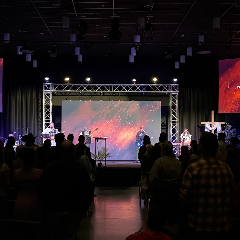 View from the audience of a worship band playing music on a brightly lit stage in a dark hall. Large screens on stage show lyrics for the congregation.