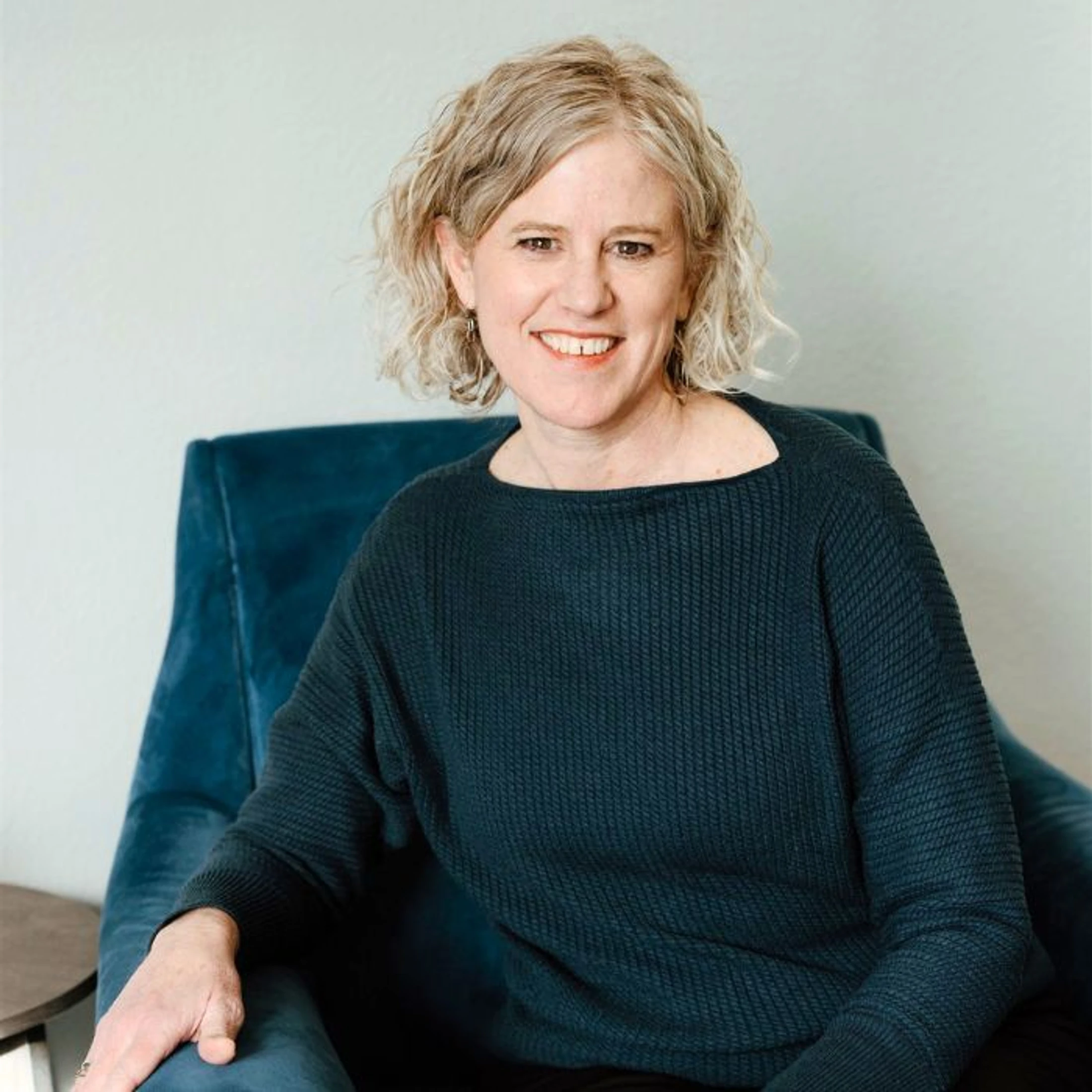 A smiling woman with short, wavy, light-colored hair sits in a blue armchair. She is wearing a dark, long-sleeved knit sweater and looking at the camera.