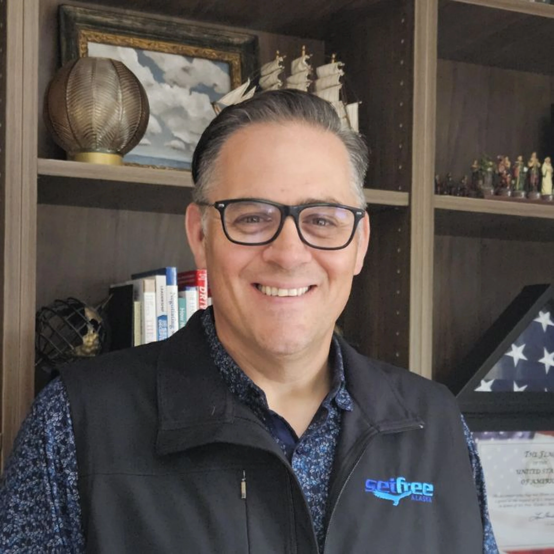 A smiling, middle-aged man with glasses wears a black vest over a patterned shirt, posing for a headshot in front of a bookshelf in his office.