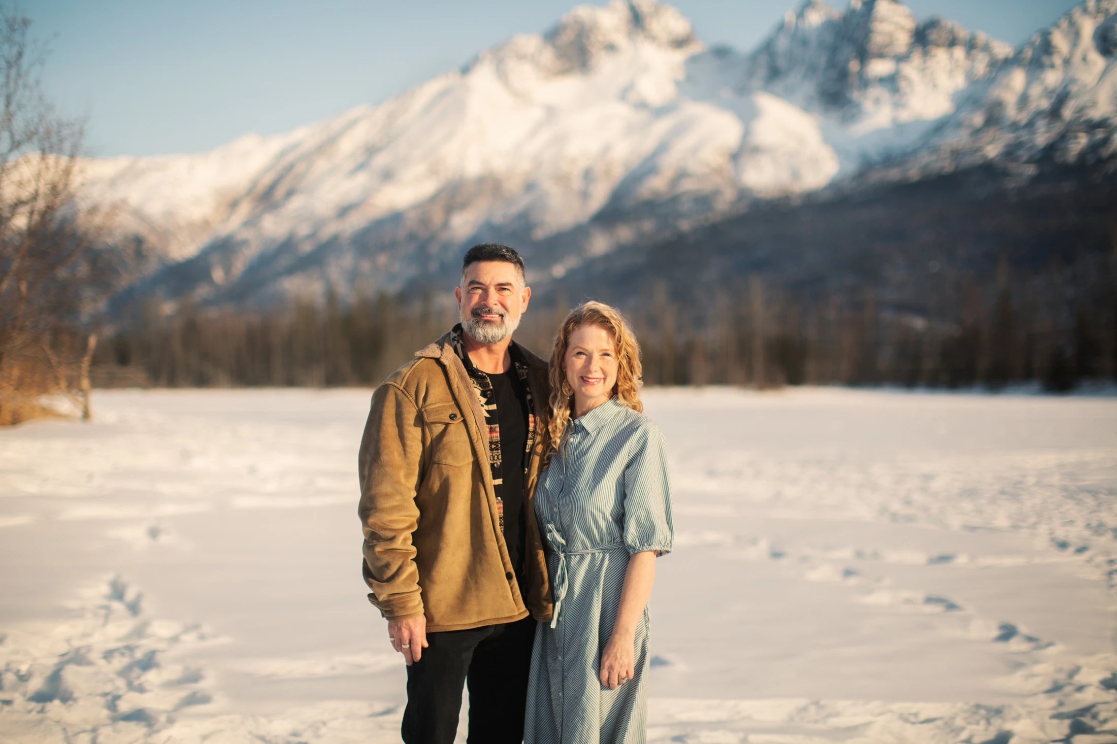 A smiling man and woman stand close together in a snowy field, with large, out-of-focus, snow-covered mountains in the background under a clear sky.