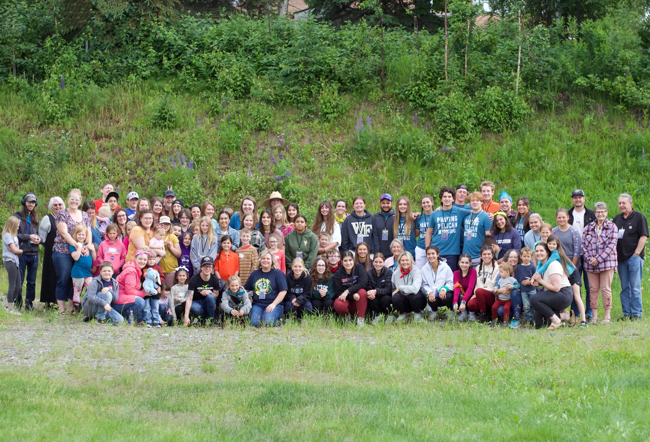 A large, diverse group of smiling people of all ages posing for a photograph on a grassy lawn in front of a hill covered in green trees and bushes.