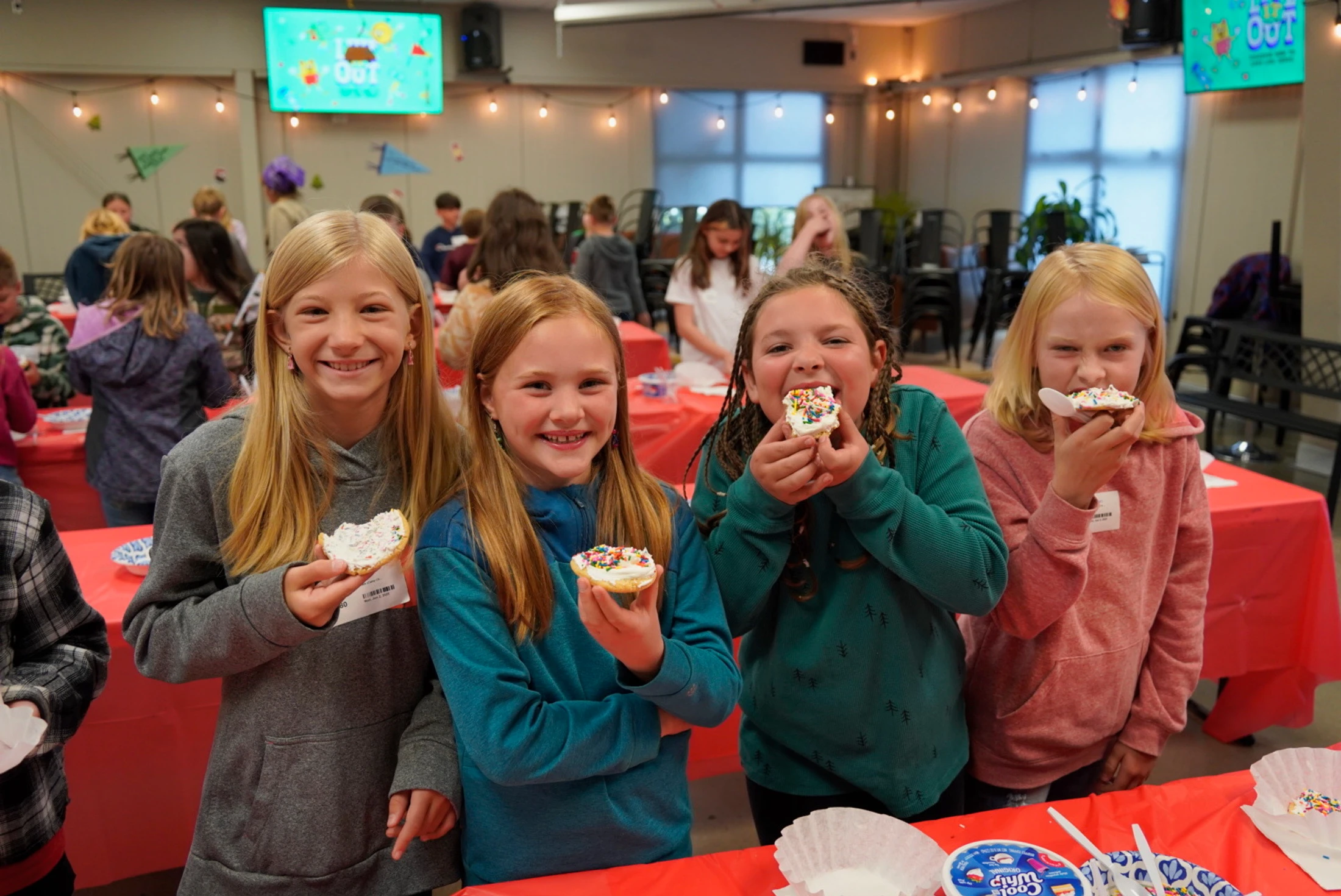 Four young girls in hoodies smile while holding and eating cookies with white frosting and colorful sprinkles at a party with red tablecloths.