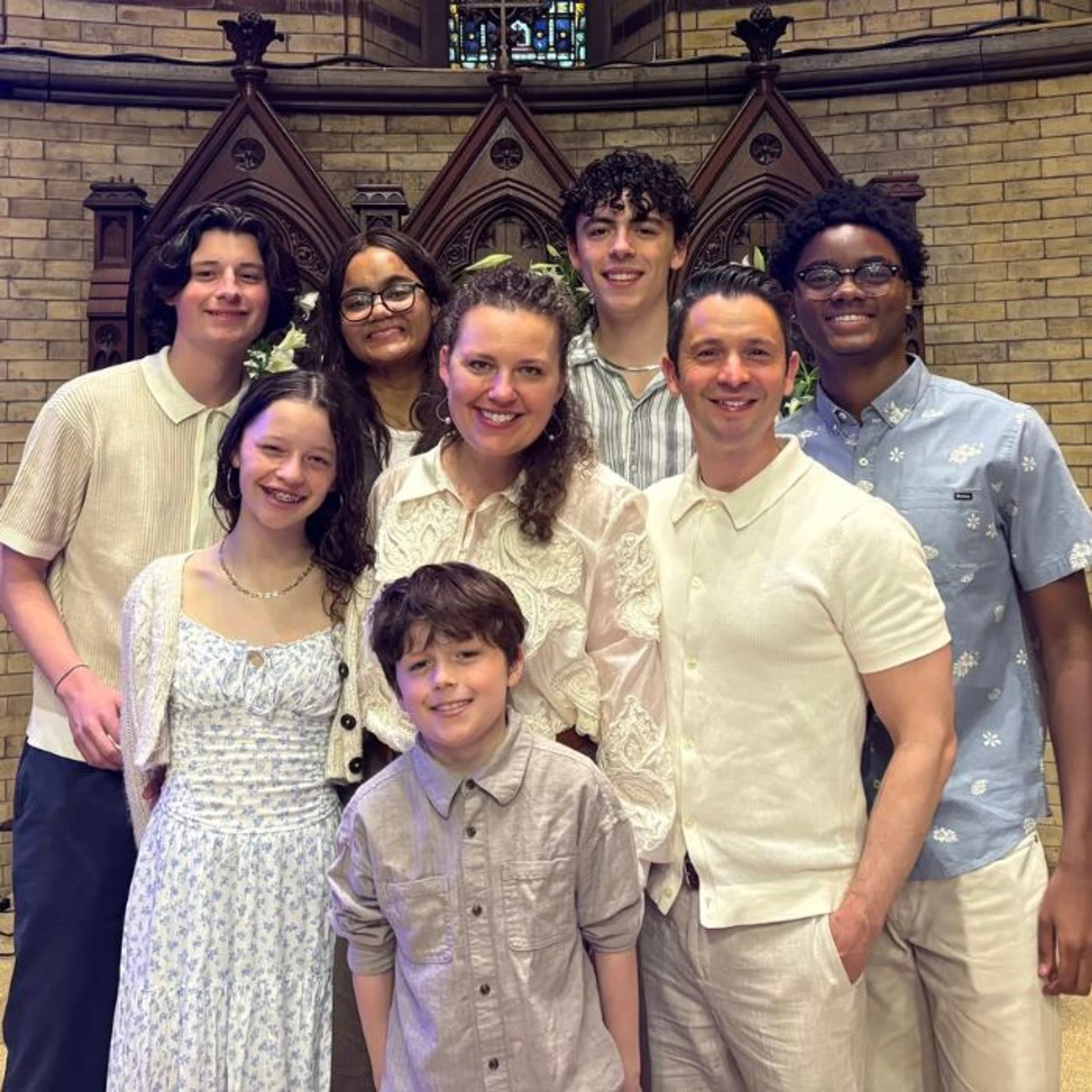 A smiling family of eight, two adults and six children, pose for a group photo inside a church. They are dressed in light-colored clothing.