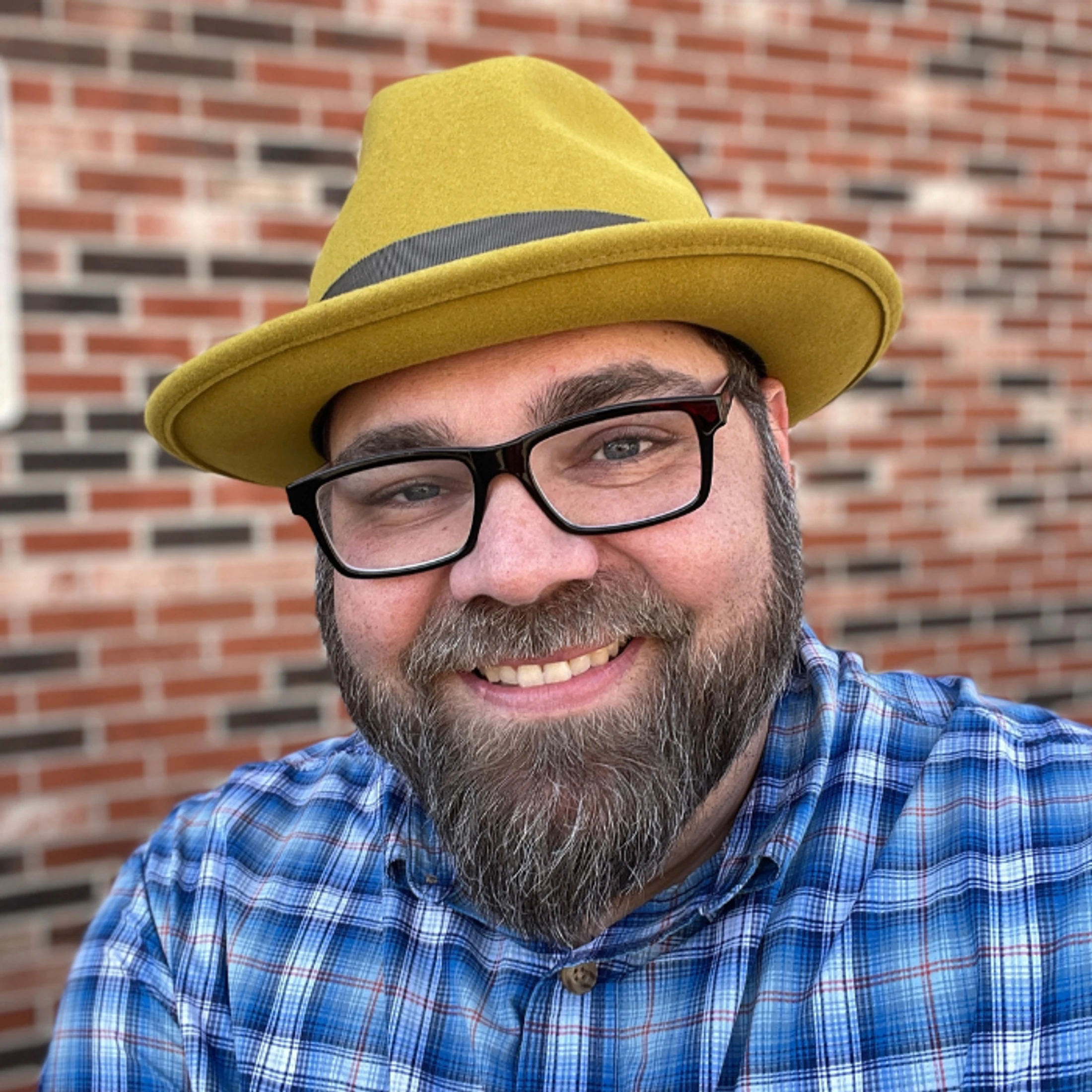 A close-up portrait of a smiling man with a beard and glasses. He is wearing a yellow fedora hat and a blue plaid shirt, with a brick wall in the background.