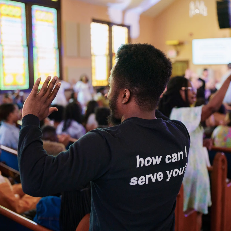 A man seen from behind in a black shirt reading 'how can I serve you?' raises his hand in worship during a church service surrounded by the congregation.