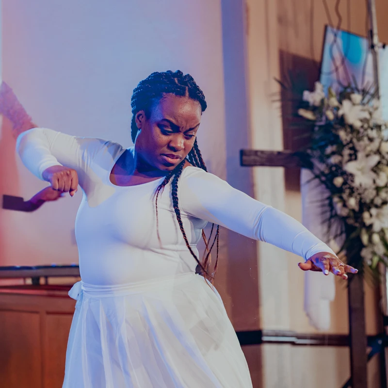 A Black woman with long braids, dressed in all white, performs an emotional dance in a church. A wooden cross and white flowers are in the background.