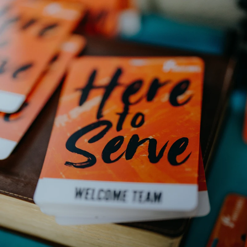 A close-up of a stack of orange cards with 'Here to Serve' and 'Welcome Team' printed in black font. The cards sit on a brown leather-like surface.