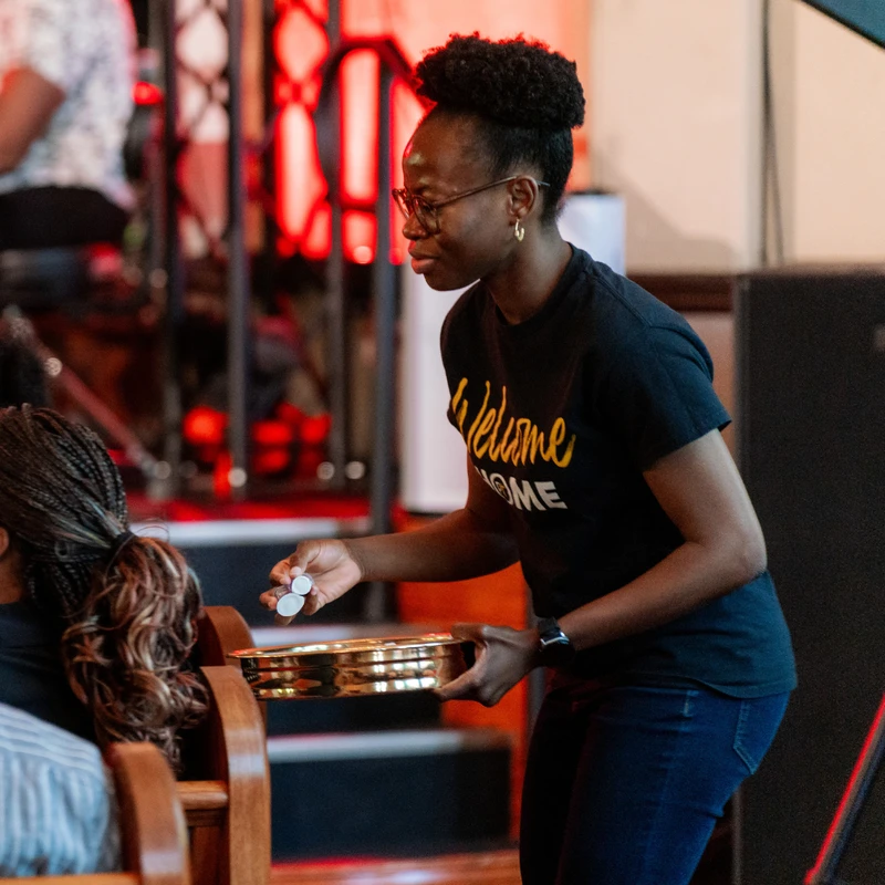 A woman in a black t-shirt that says 'Welcome Home' holds a brass communion plate and serves elements to a person in a church pew.