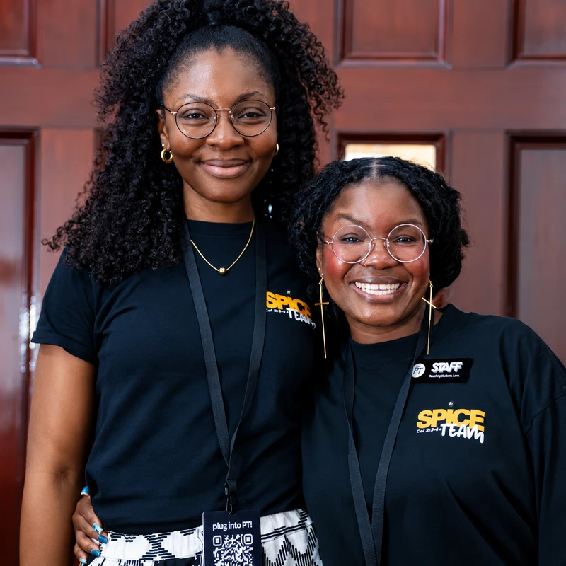 Two Black women wearing matching black 'Spice Team' staff shirts and glasses smile for the camera while standing in front of a dark wooden door.