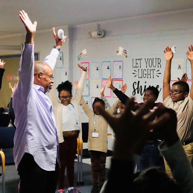 An adult male instructor and a group of diverse, smiling children stand in a circle with their hands raised joyfully in a classroom setting.