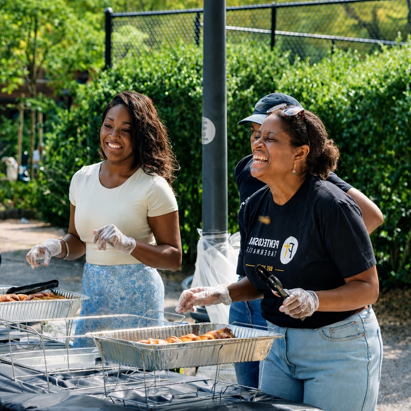 Two Black women volunteers smile and laugh while serving food from foil trays at a sunny outdoor cookout. One woman holds tongs and laughs heartily.