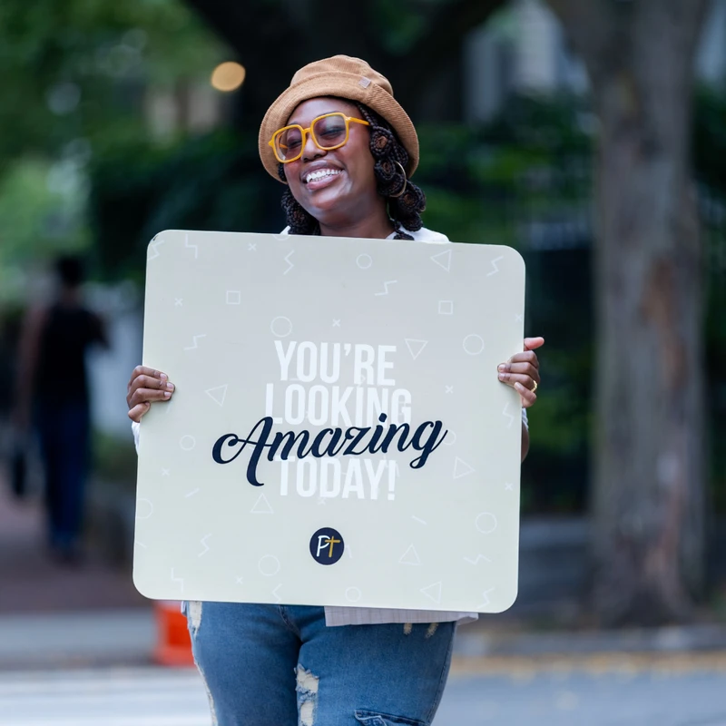 A smiling woman with a brown hat and orange glasses holds a sign that reads, "You're Looking Amazing Today!" on a city street.