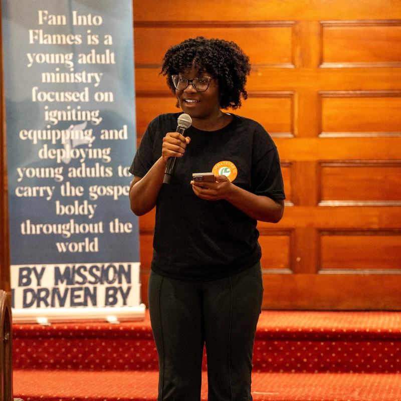 A young Black woman with glasses smiles while speaking into a microphone. She stands on a red carpet next to a blue banner for a young adult ministry.