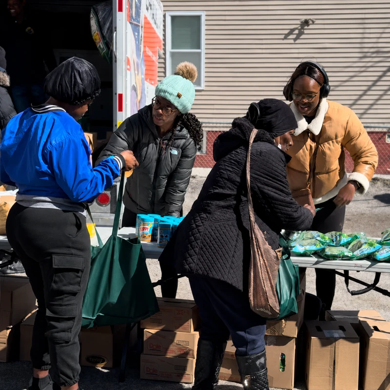Four women stand around a table with donated groceries like peanut butter and fresh greens during an outdoor food distribution event next to a truck.