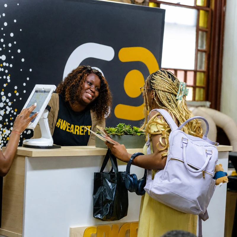 A smiling woman wearing a #WEAREPT t-shirt greets another woman with blonde braids at a registration desk, while a third person uses a check-in tablet.