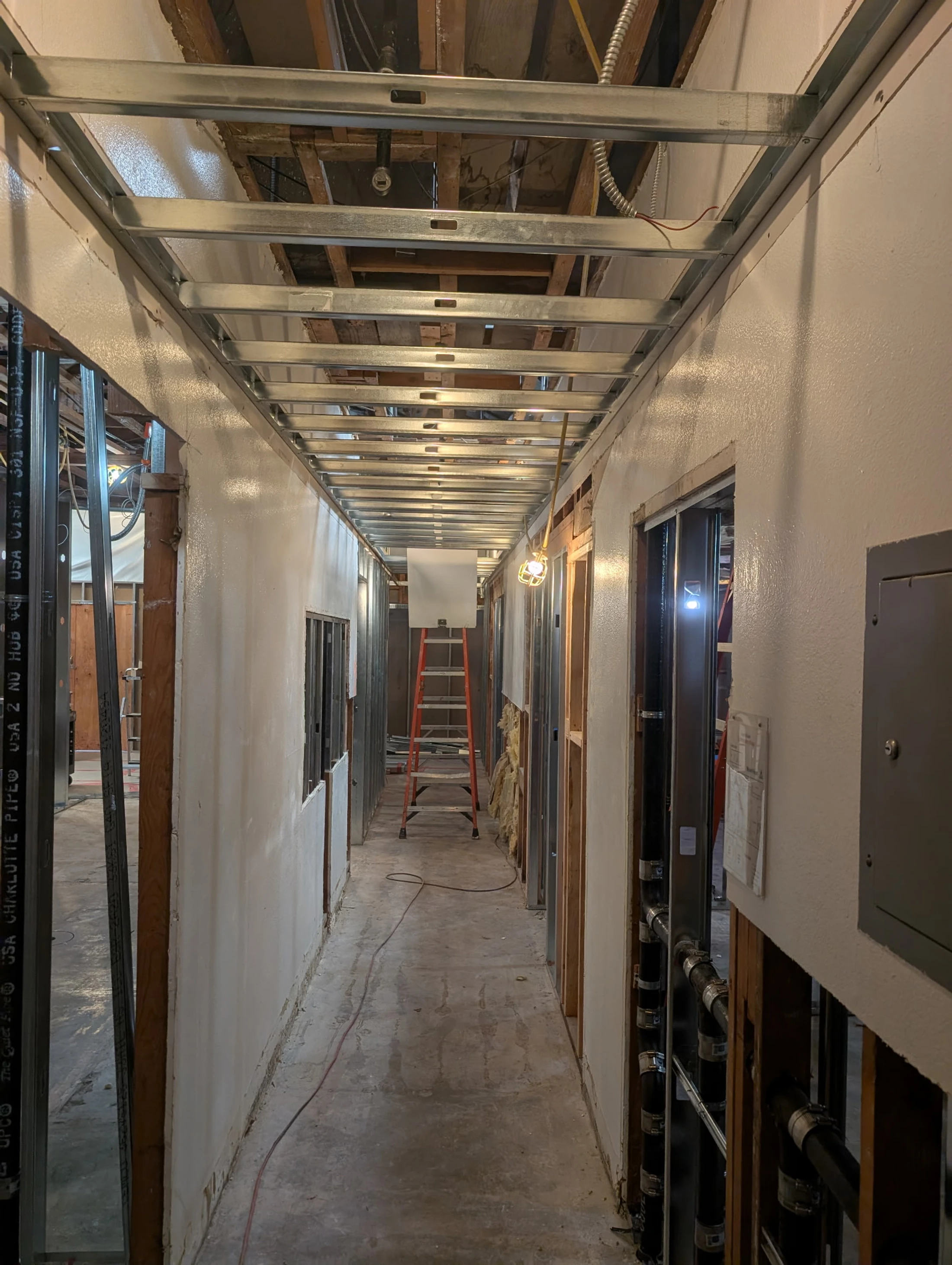 A long hallway during renovation with an exposed metal ceiling grid, unfinished walls with visible framing and pipes, and a concrete floor.