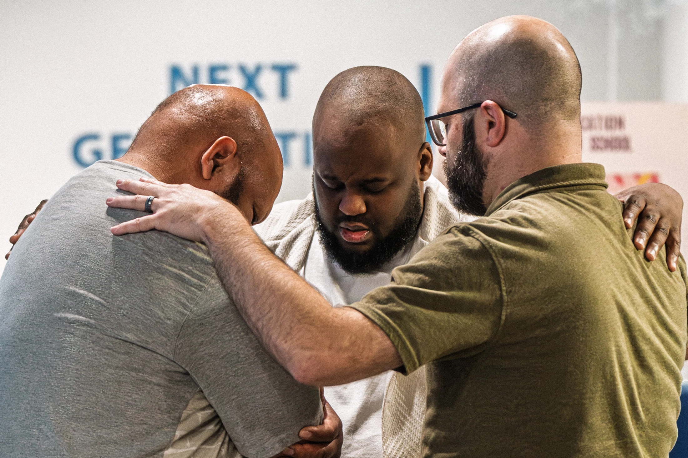 Three diverse men are huddled together with their arms around each other, heads bowed and eyes closed in a moment of emotional prayer.