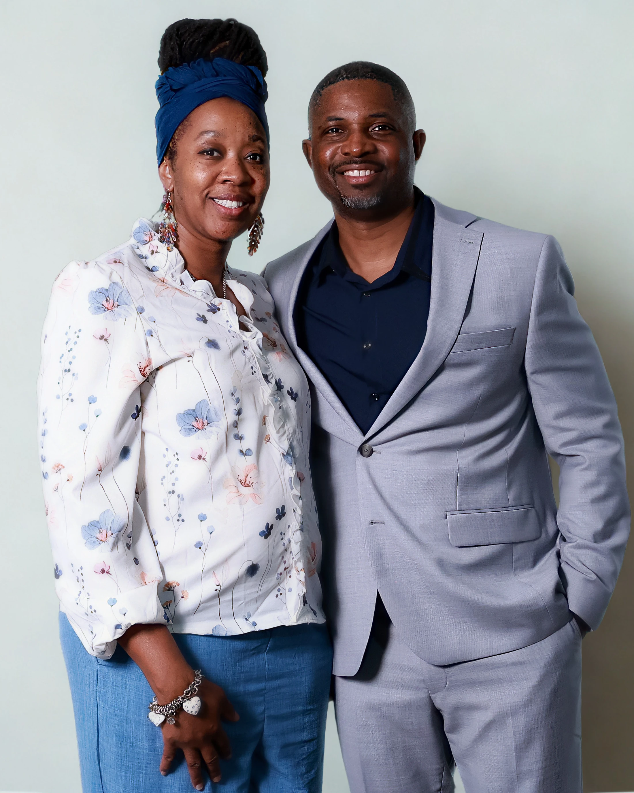 A smiling Black couple stands together against a light background. The woman wears a floral blouse and a blue headwrap, and the man wears a light gray suit.